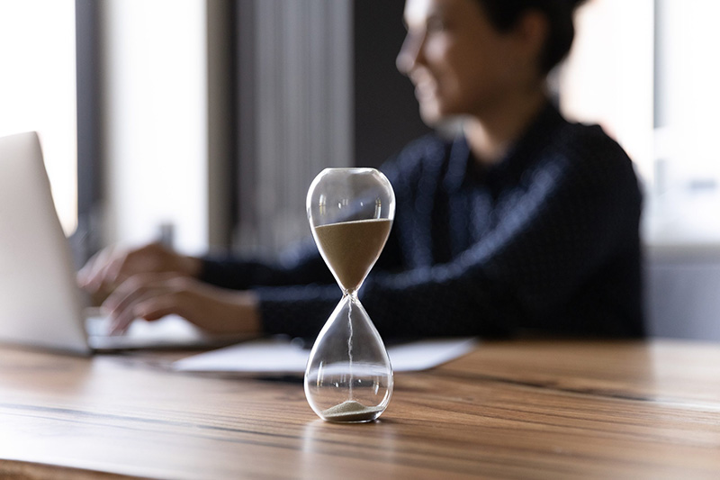 A glass hourglass with sand running through it sits on a wooden desk, while a person works on a laptop in the blurred background.