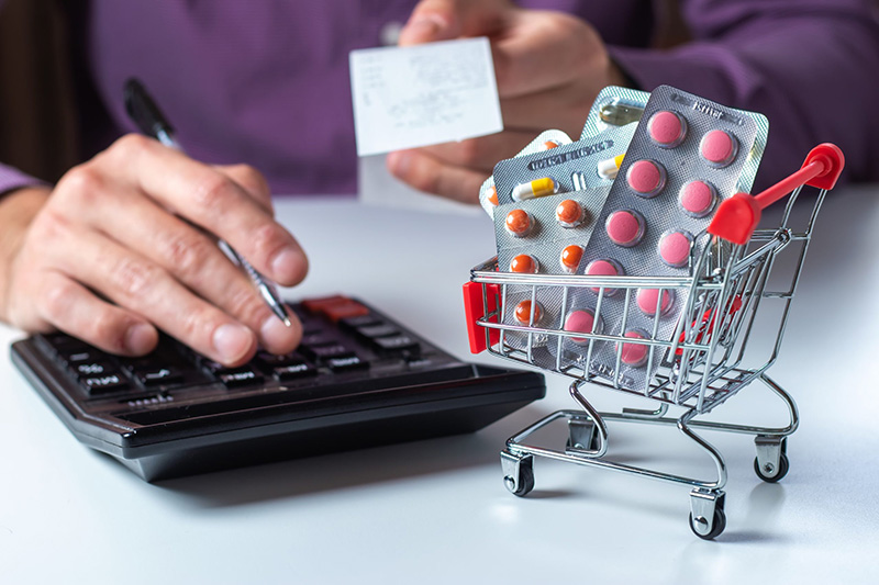 A person uses a calculator and holds a receipt while a small shopping cart filled with blister packs of pills sits on the table, symbolizing the cost of medication.