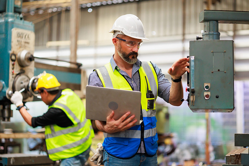 A man in a hard hat and safety vest holds a laptop while inspecting industrial machinery in a factory. Another worker in the background operates equipment, both focused on their tasks in a busy industrial setting.
