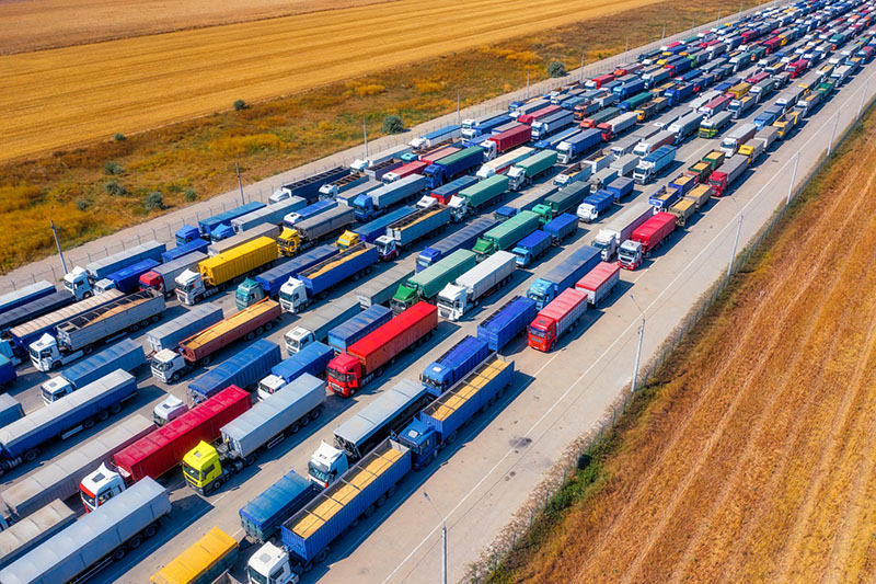 Aerial view of a long line of colorful cargo trucks parked in multiple lanes on a wide road next to agricultural fields, suggesting a traffic jam or border checkpoint.
