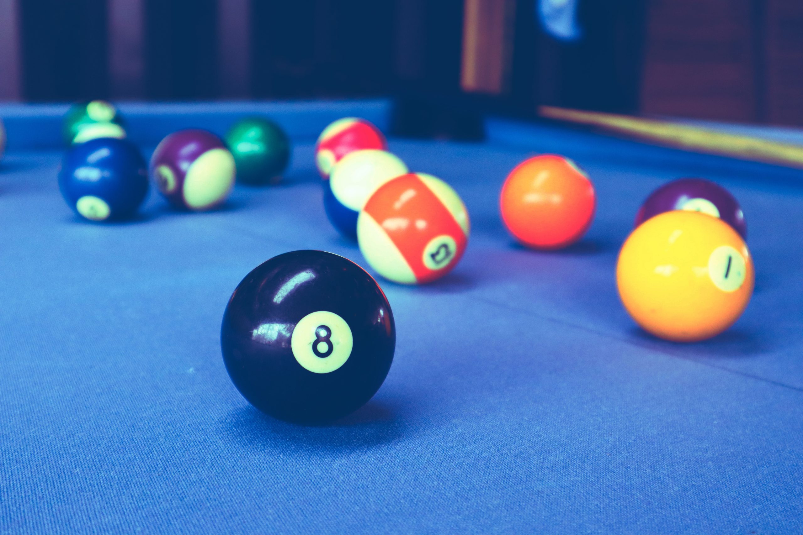 Several billiard balls, including the black 8 ball in the foreground, are scattered across a blue pool table. The image is brightly lit and shows other colored balls out of focus in the background.