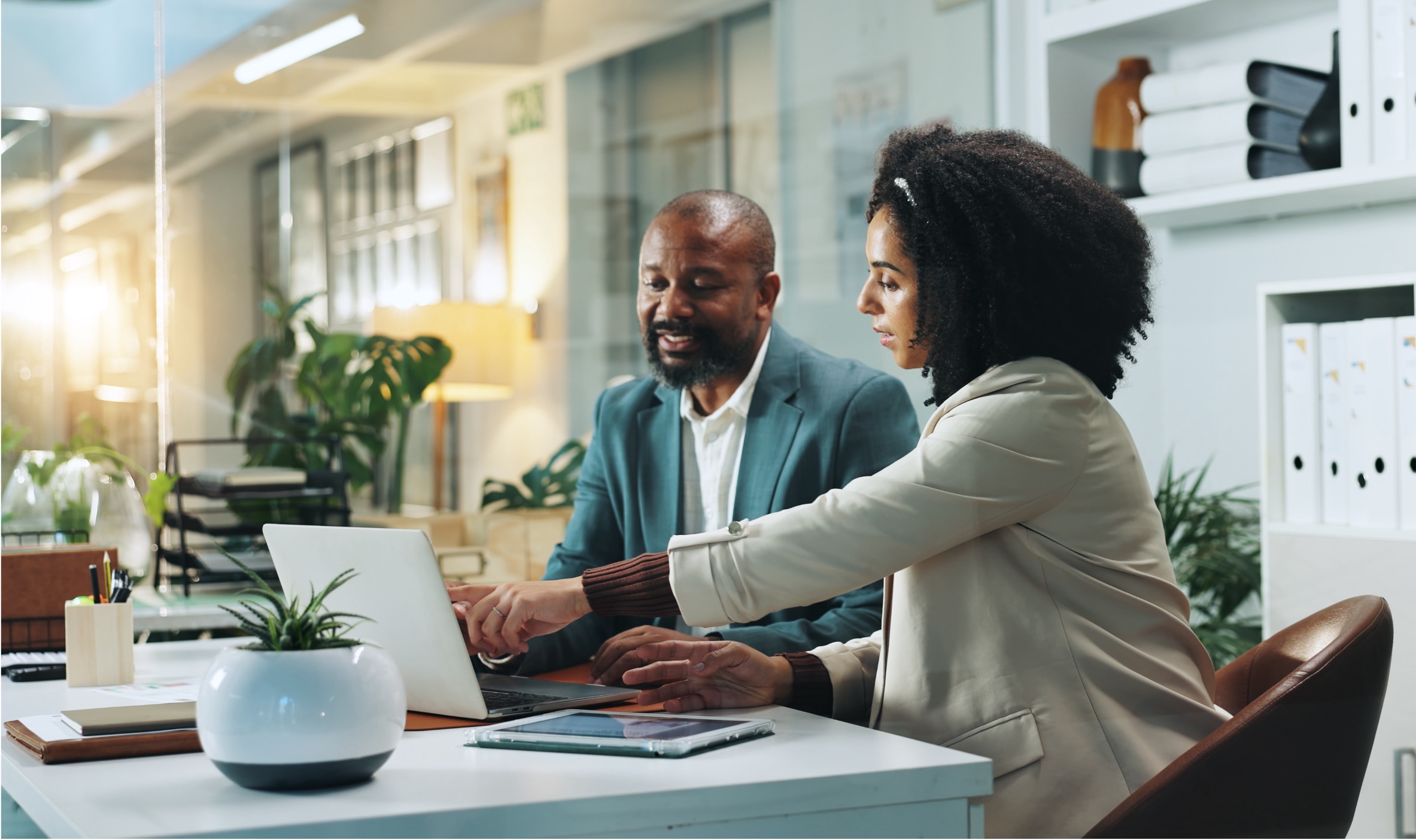 Two professionals sit at a desk in a modern office, collaborating while looking at a laptop. A potted plant, documents, and office supplies are on the desk, with shelves and large windows in the background.
